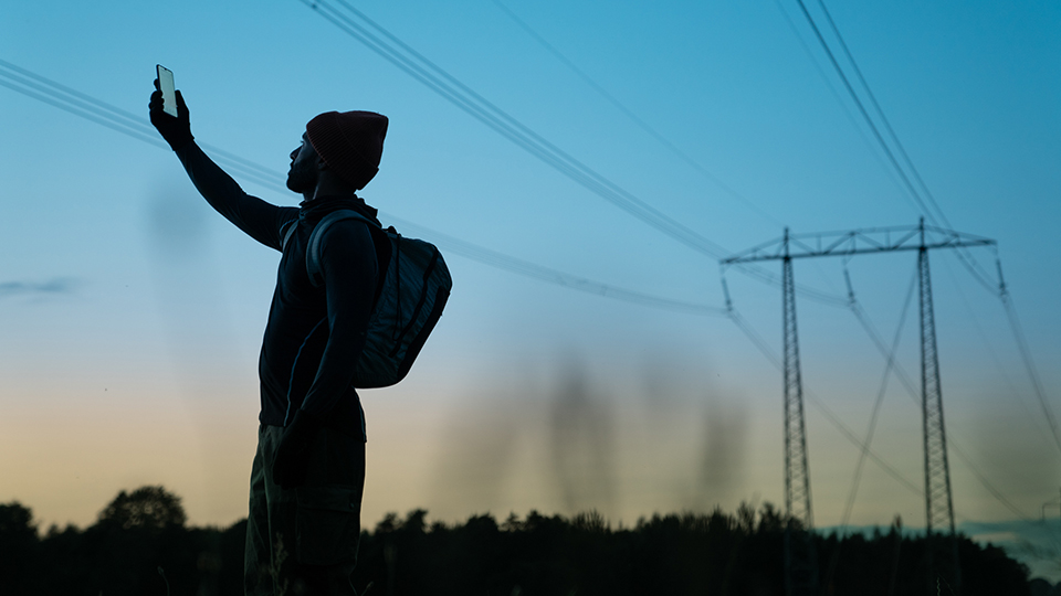 Ett foto på en person med  mössa står vid en kraftledning och håller upp en mobiltelefon mot himlen.
