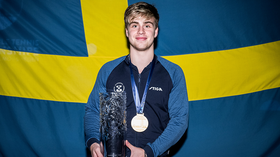 Bordtennisspelaren och kommunambassadören Truls Möregårdh har en guldmedalj runt halsen och han håller en glaspokal i händerna. I bakgrunden syns en svensk flagga.