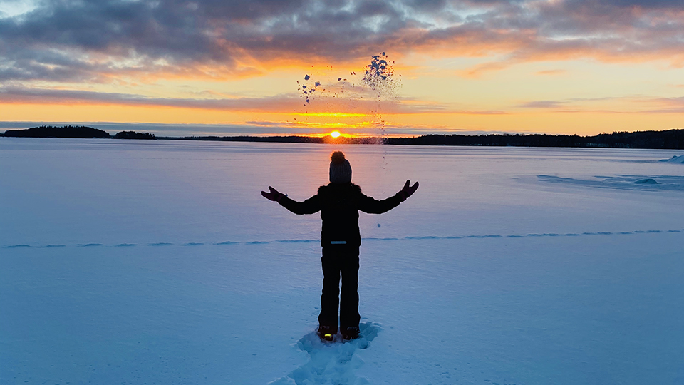 Foto på ett barn som kastar upp snö medan solen går ner vid horisonten.